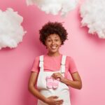 Beautiful dark skinned expectant mother dressed in white sarafan holds babys socks over belly and enjoys moment of maternity poses against pink background with white clouds. Pregnancy concept