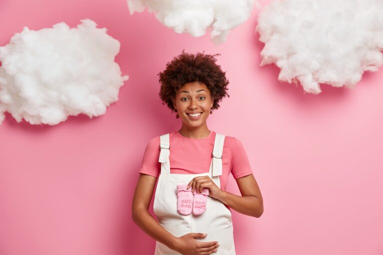 Beautiful dark skinned expectant mother dressed in white sarafan holds babys socks over belly and enjoys moment of maternity poses against pink background with white clouds. Pregnancy concept